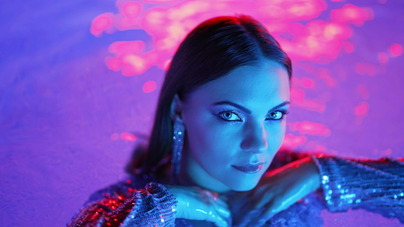 Comment s’habiller avec élégance pour une soirée chic ? Beautiful woman posing in pool water under neon color light. Party, attractive chic in shining dress