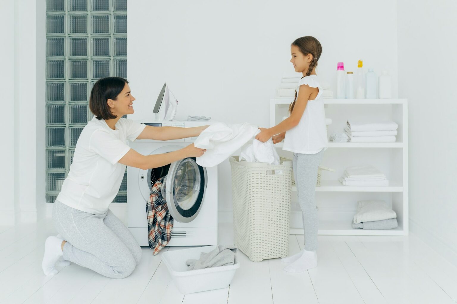 Happy mother loads clothes in washing machine, little girl helps, gives white linen from basket